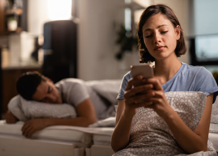 young woman using smart phone in bedroom while her boyfriend is sleeping